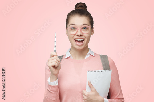 Portrait of young female showing idea gesture with her pen to indicate significant information with smile and eyes wide open, isolated on pink background