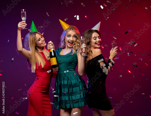 Portrait of three amazing, cute girls in fashion dresses and party caps, holding glass with champagne, celebrating the New year or Birthday party, standing over colorful background