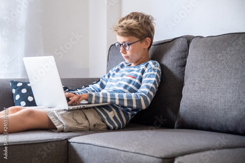 Beautiful blond child sitting on the couch at home using computer. Young boy relaxing with laptop free time online games. Teen working and chat with internet friends. Student doing homework on the web