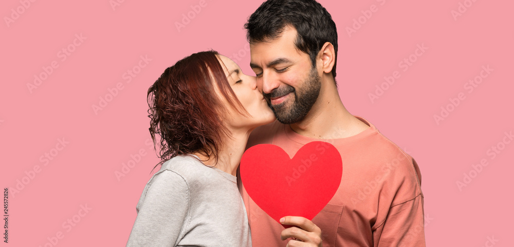 Couple in valentine day holding a heart symbol and kissing over isolated pink background