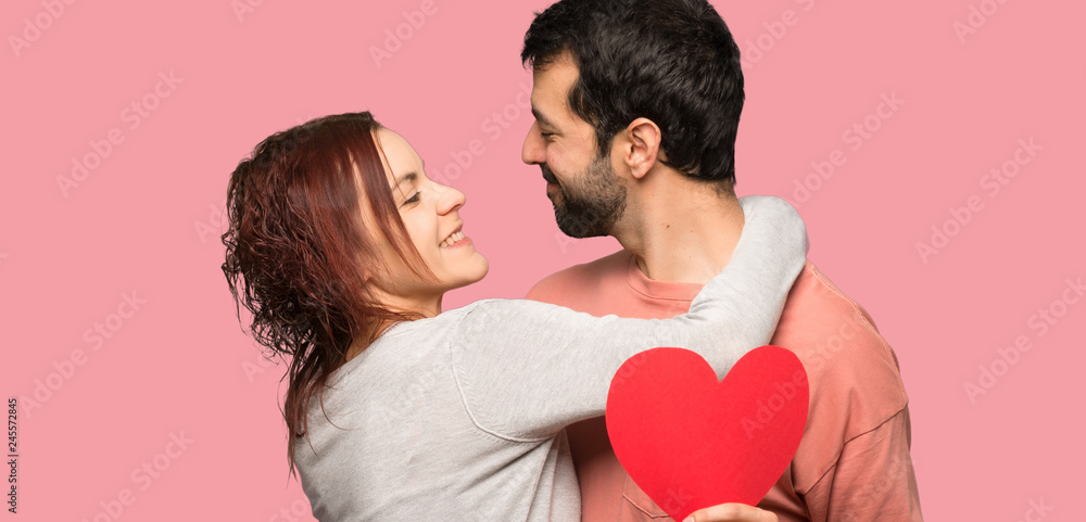 Couple in valentine day holding a heart symbol and kissing over isolated pink background Couple in valentine day holding a heart symbol and kissing over isolated pink background