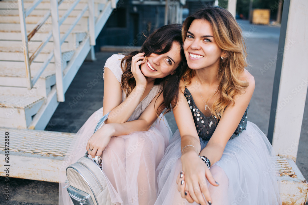 Portrait of two attractive girls in tulle skirts sitting outdoor on stairs. They smiling to camera