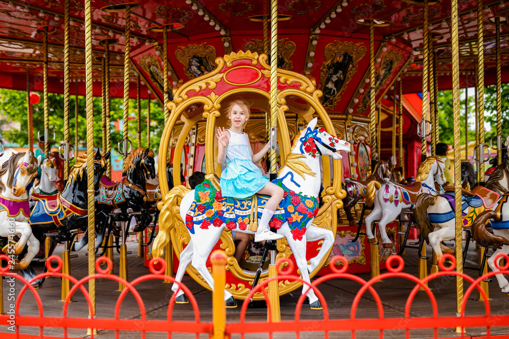 Blonde girl with two braids in white and blue dress riding colorful horse in the merry-go-round carousel in the entertainment park