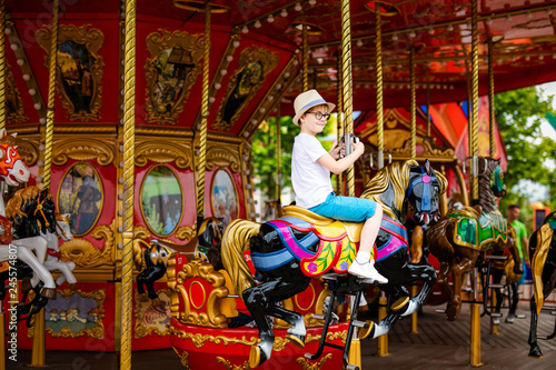 Blonde boy in the straw hat and big glasses riding colorful horse in the merry-go-round carousel in the entertainment park © Lena May