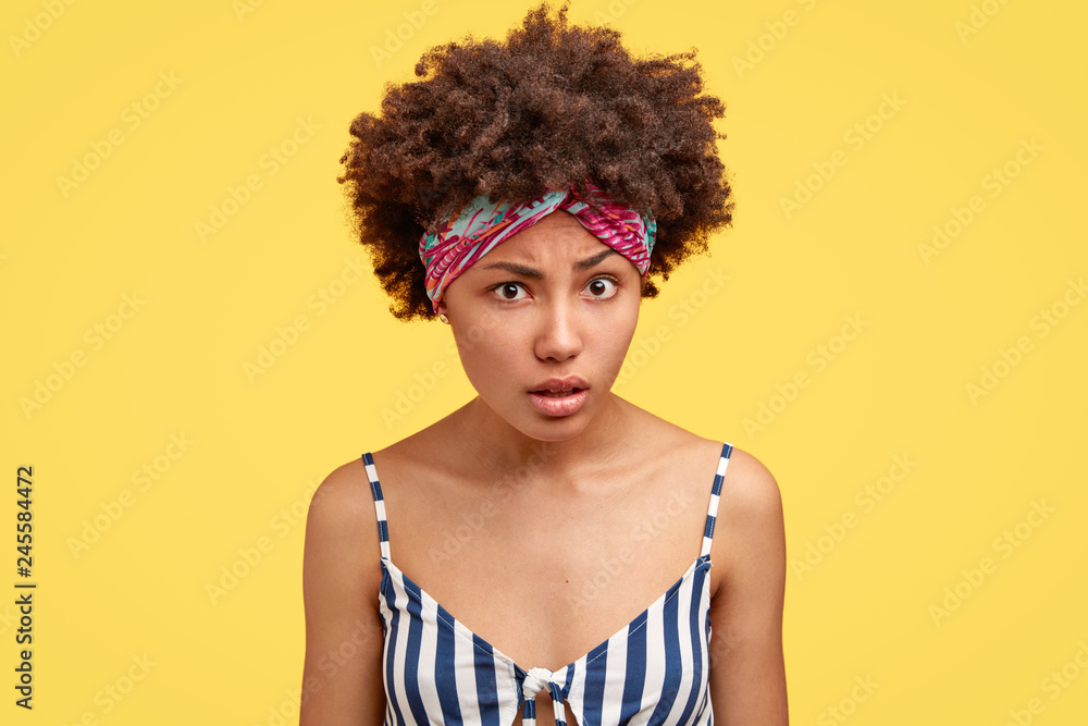 Pretty young woman with crisp hair, wears headband and striped top, looks with indignation, tries to understand something, models against yellow bckground. People, reaction and facial expressions Pretty young woman with crisp hair, wears headband and striped top, looks with indignation, tries to understand something, models against yellow bckground. People, reaction and facial expressions