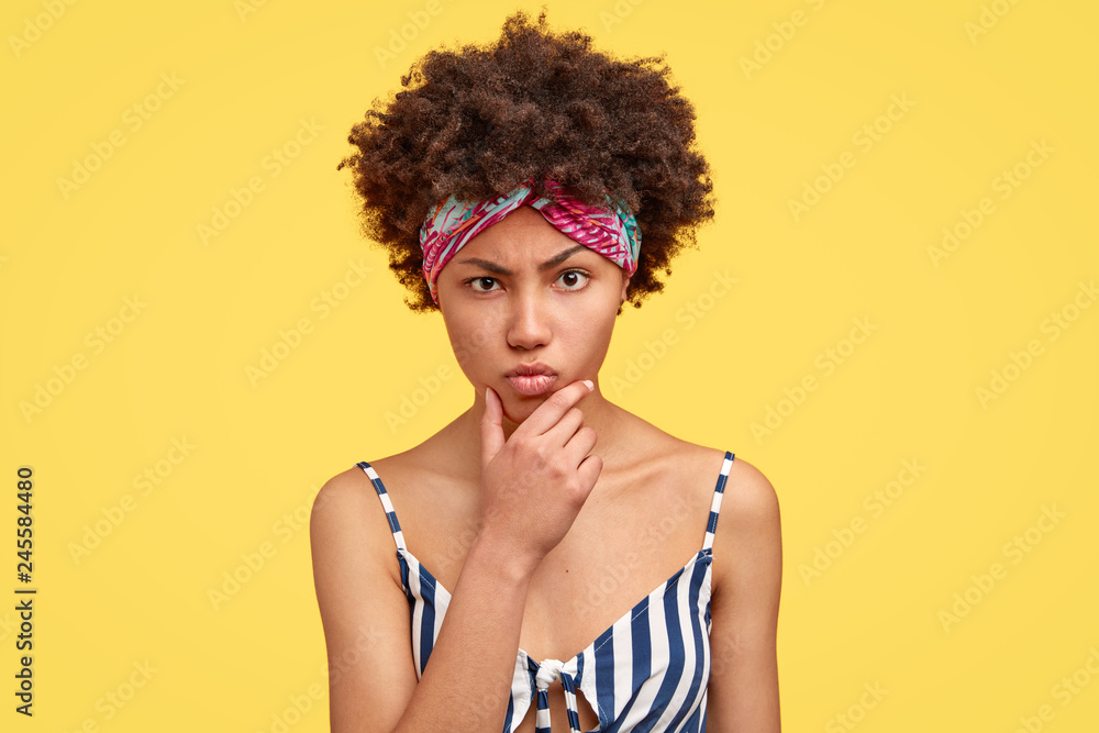 Displeased angry dark skinned young woman holds chin, claims about something bad, has sullen expression, crisp hair, wears headband, models against yellow background. Negative emotions concept Displeased angry dark skinned young woman holds chin, claims about something bad, has sullen expression, crisp hair, wears headband, models against yellow background. Negative emotions concept