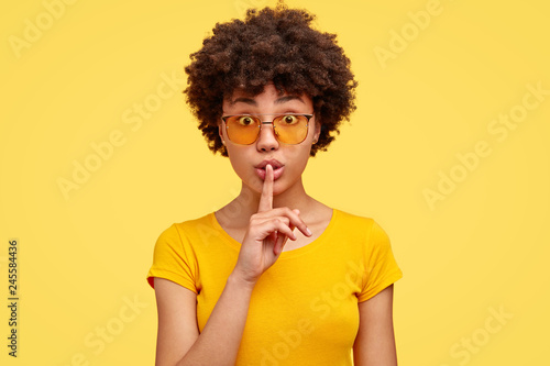 Stupefied secret dark skinned woman makes gesture quietly, asks remain silent, gossips about something, looks mysteriously as tells secret, wears bright yellow t shirt, stands indoor. Shh, dont speak photo