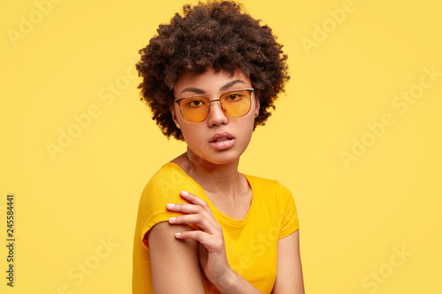 Sideways shot of calm serious emotionless woman keeps arms crossed, wears yellow t shirt and shades, has frizzy hair, being without emotions, has natural beauty. People and facial expressions photo