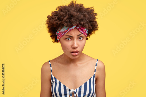 Pretty young woman with crisp hair, wears headband and striped top, looks with indignation, tries to understand something, models against yellow bckground. People, reaction and facial expressions