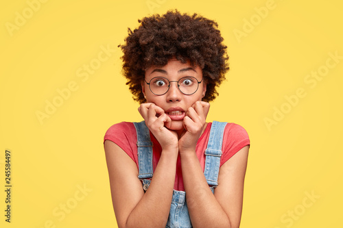 Headshot of frustrated mixed race young woman has crisp hair, bites finger nails with nervous expression, stares with eyes full of fear, wears casual clothes, stands against yellow studio wall
