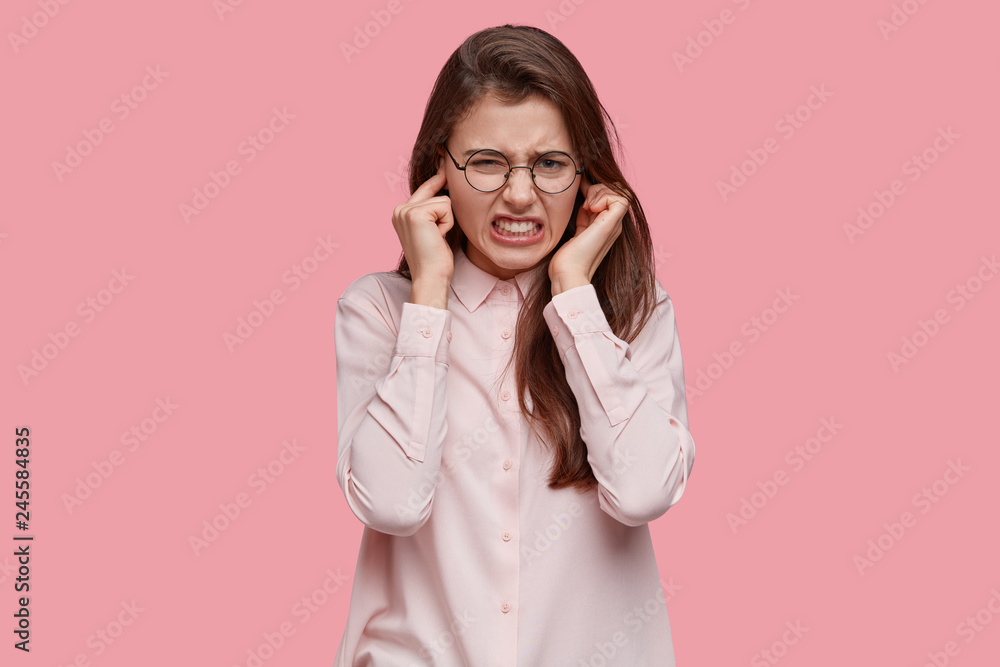 Frustrated young lady plugs ears, clenches teeth from annoyance, ignores annoying sound, wears transparent spectacles, hears noise coming from neighbours, isolated over pink background. Decibel Frustrated young lady plugs ears, clenches teeth from annoyance, ignores annoying sound, wears transparent spectacles, hears noise coming from neighbours, isolated over pink background. Decibel
