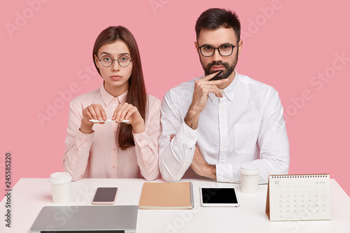 Tableau sur toile Joyful self assured woman with positive smile holds pen, serious bearded man in white shirt, collaborate together, use modern gadgets, pose at white table against pink background