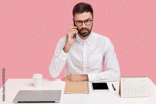 Strict angry male boss calls on digital smart phone, does remote work, consults with business partner, wears spectacles and white shirt, has mobile conversation, makes research, sits over pink wall photo