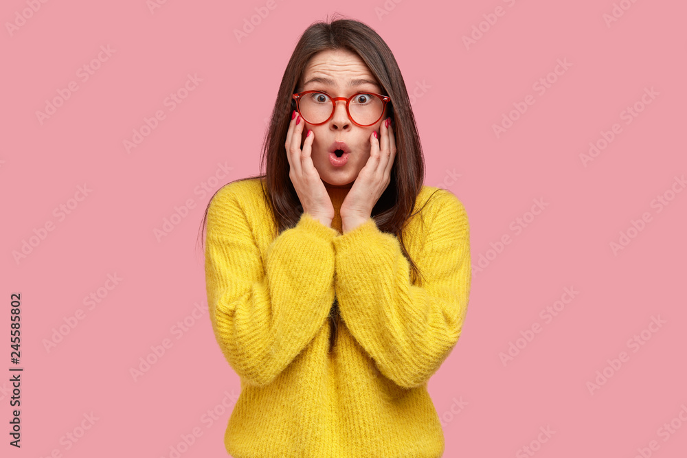 Φωτογραφία Woman in panic touches cheeks with both palms, can not believe her eyes, wonders while hears rumors, dressed in yellow sweater, isolated over pink background, sees no way