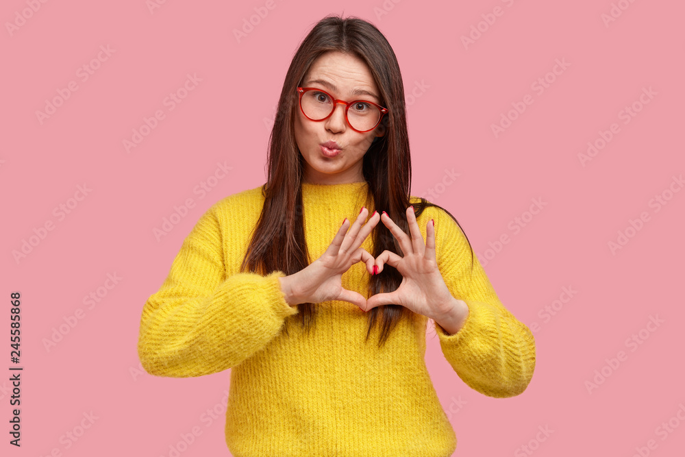 Φωτογραφία Romantic pleased young woman makes heart gesture, wears yellow sweater, transparent glasses, pouts lips, expresses love, has charming look, isolated over pink background