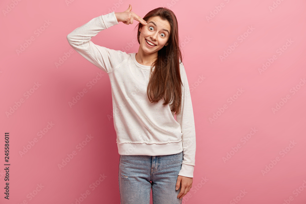 Happy young woman shoots with fingers in temple, makes suicide gesture, smiles broadly, tilts head, tries kill herself, wears jumper and jeans, isolated over pink background, fed up with something Happy young woman shoots with fingers in temple, makes suicide gesture, smiles broadly, tilts head, tries kill herself, wears jumper and jeans, isolated over pink background, fed up with something