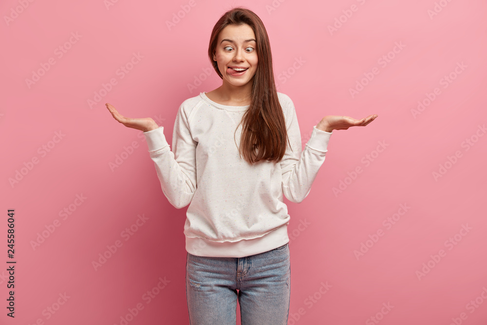 Crazy funny European girl sticks out tongue and crosses eyes, spreads palms, holds copy space, makes grimace, foolishes indoor, dressed in casual jumper and jeans, isolated over pink background Crazy funny European girl sticks out tongue and crosses eyes, spreads palms, holds copy space, makes grimace, foolishes indoor, dressed in casual jumper and jeans, isolated over pink background