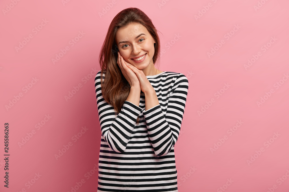 Studio shot of attractive woman with healthy skin, pleasant appearance, keeps hands together near cheek, has dark hair, smiles gently, wears casual striped clothes, isolated over pink background Studio shot of attractive woman with healthy skin, pleasant appearance, keeps hands together near cheek, has dark hair, smiles gently, wears casual striped clothes, isolated over pink background