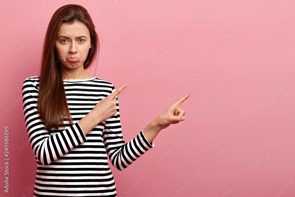 Distressed European woman purses lips, looks with displeasure, dressed in striped clothes, shows copy space for advertising content, feels dejected, displeased, isolated over pink background Distressed European woman purses lips, looks with displeasure, dressed in striped clothes, shows copy space for advertising content, feels dejected, displeased, isolated over pink background