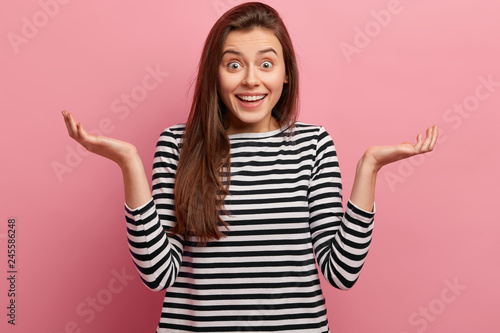 Clueless cheerful woman with dark long hair, has opened eyes, spreads palms, wears striped black and white jumper, feels uncertainty, isolated over pink background. People and attitude concept