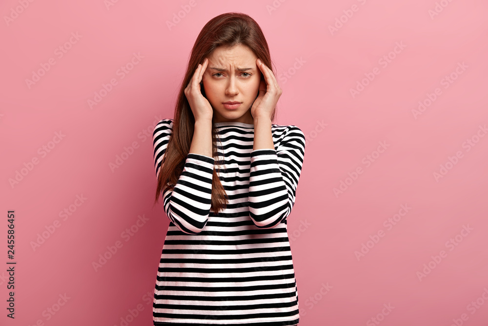 Beautiful woman keeps hands on temples, frowns face with pain, suffers from headache, annoyed with noisy sounds, wears casual striped jumper, models against pink studio wall. Sickness concept Beautiful woman keeps hands on temples, frowns face with pain, suffers from headache, annoyed with noisy sounds, wears casual striped jumper, models against pink studio wall. Sickness concept