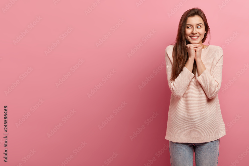 Shot of attractive woman keeps hands together near face, focused aside, smiles broadly, shows white teeth, wears casual jumper and jeans, isolated over pink studio wall, empty space for your slogan Shot of attractive woman keeps hands together near face, focused aside, smiles broadly, shows white teeth, wears casual jumper and jeans, isolated over pink studio wall, empty space for your slogan