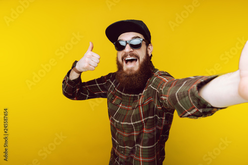 Like ! Bearded guy taking selfie in black hat and sunglasses and gesturing thumb up, good, like symbol