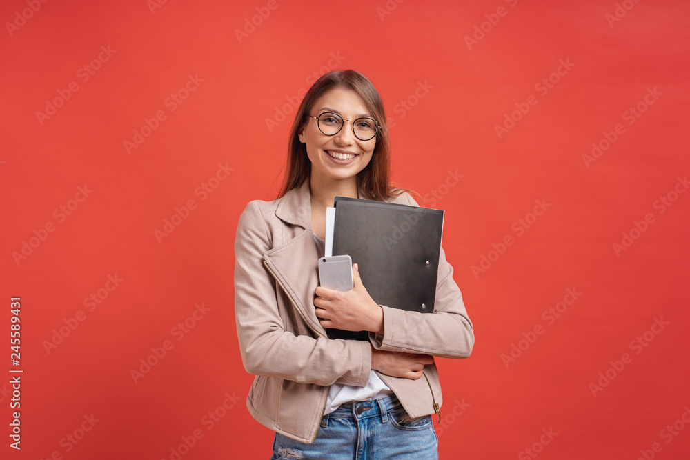 Young smiling student or intern in eyeglasses standing with a folder on red background. - obrazy, fototapety, plakaty 