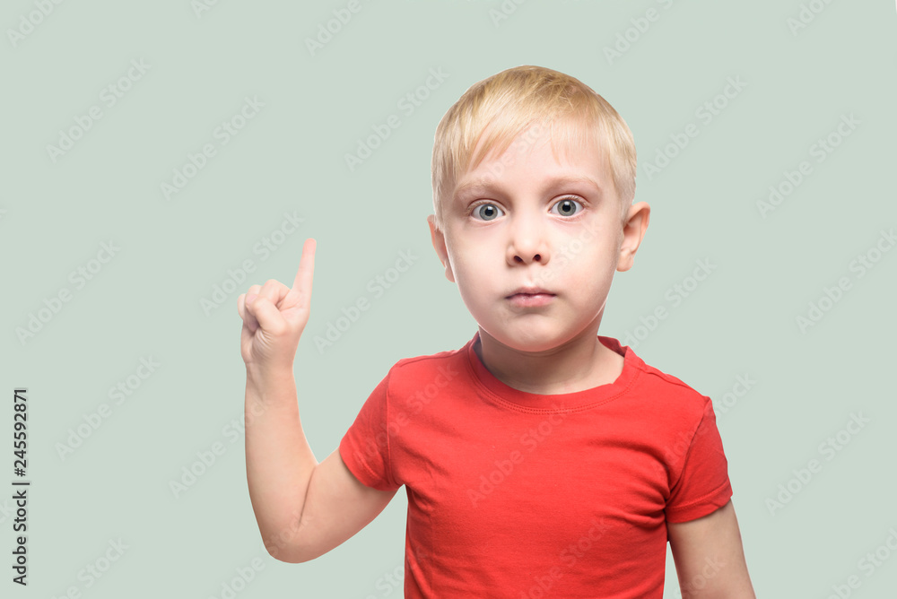 Surprised little blond boy in red t-shirt stands and points with index finger upwards. Isolate on light green bacground Surprised little blond boy in red t-shirt stands and points with index finger upwards. Isolate on light green bacground