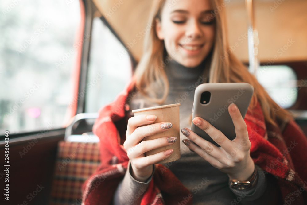 Fototapeta premium Happy girl uses a smartphone and drinks coffee in a cozy cafe. Close-up of a smartphone and a cup of coffee in their hands. Background. Copyspace