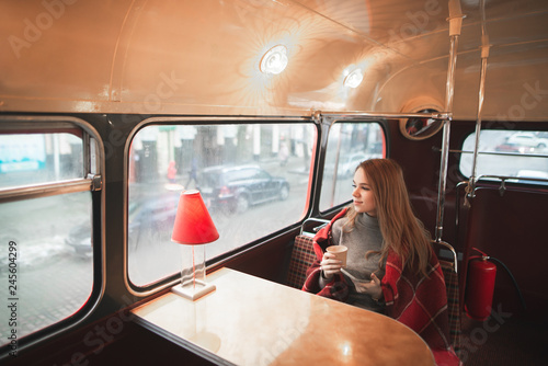 Young attractive woman sitting at the table in the bus cafe is covered with a blanket, holds a cup of coffee in her hands and looks in the window. Beautiful girl sits in a cozy original cafe.