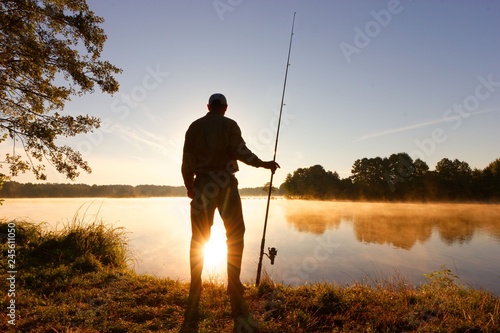 Silhouette of angler standing on the lake shore during misty sunrise