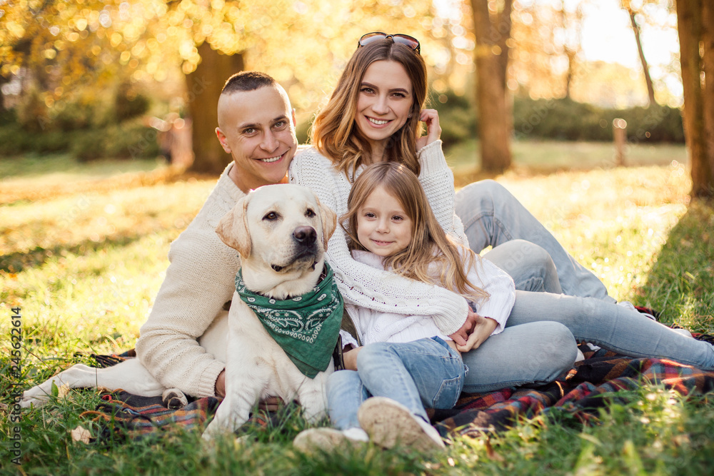 Fototapeta premium Happy beautiful family with dog labrador is having fun are sitting on green grass in park.