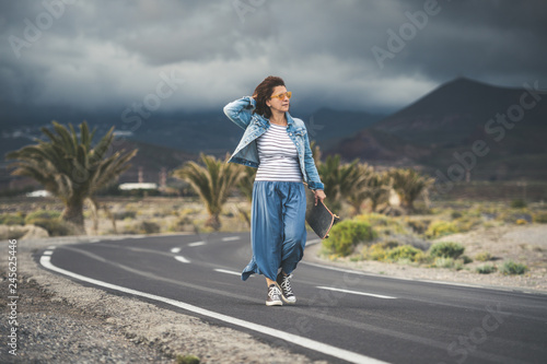 Middle aged woman skateboard smiles satisfied holding her dark hair with hand curved road with thunderstorm horizon striped knit long skirt sneakers wind free time summer holidays sunset sport