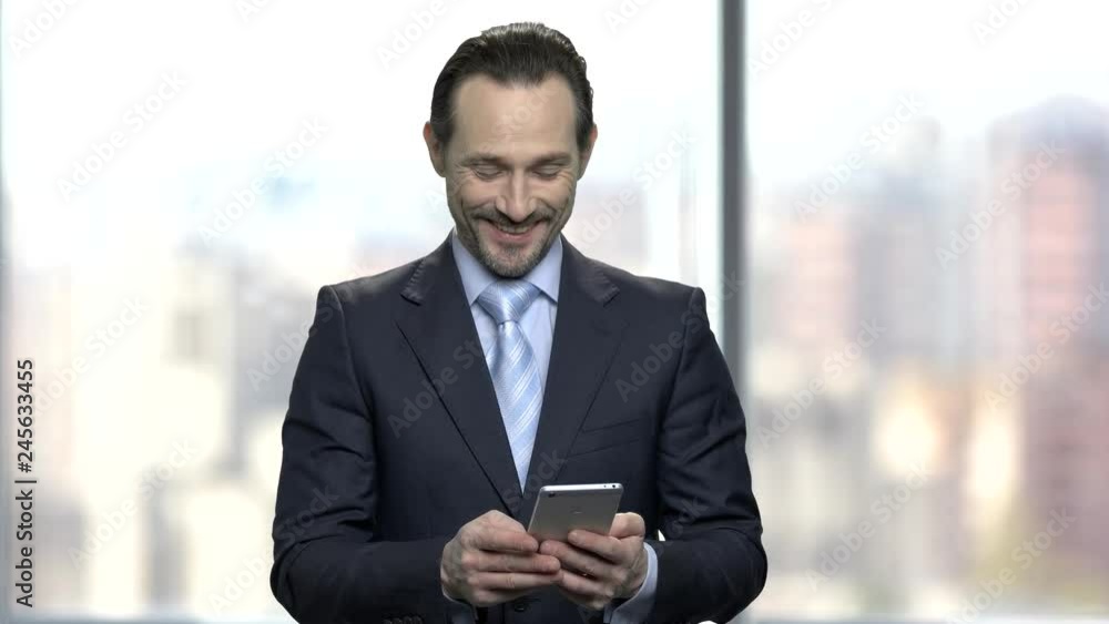 Smiling businessman reading message on his smartphone. Attractive entrepreneur using mobile phone on blurred background. Smiling businessman reading message on his smartphone. Attractive entrepreneur using mobile phone on blurred background.