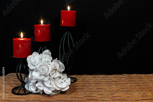 Beautiful bouquie of white roses, red candles perched on black candle holders on mesh place mat and wooden table with card and dark background. 