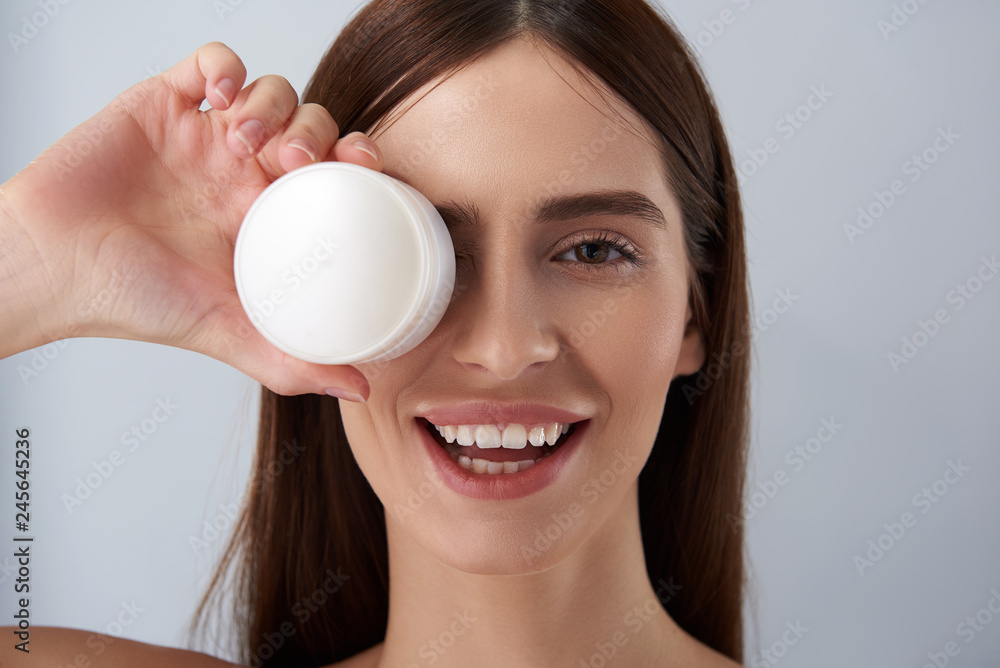 Charming brunette girl covering eye with white jar of cosmetic cream Charming brunette girl covering eye with white jar of cosmetic cream