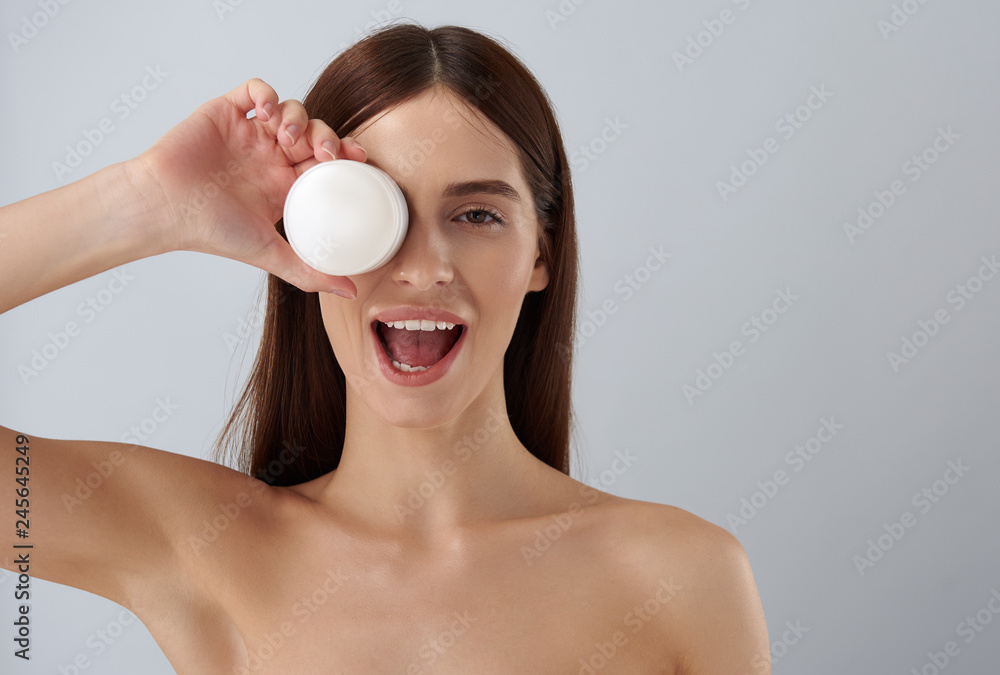 Cheerful brunette girl covering eye with white jar of cosmetic cream Cheerful brunette girl covering eye with white jar of cosmetic cream