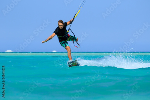 Kite surfing man surf sportsman with kite in sky on board in blue sea riding waves with water splash. Recreational activity, water sports, action, hobby and fun in summer time. Kiteboarding sport