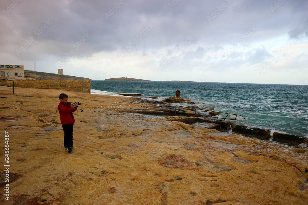 Little tourist boy make a photo of stormy mediterranean sea on Malta coast