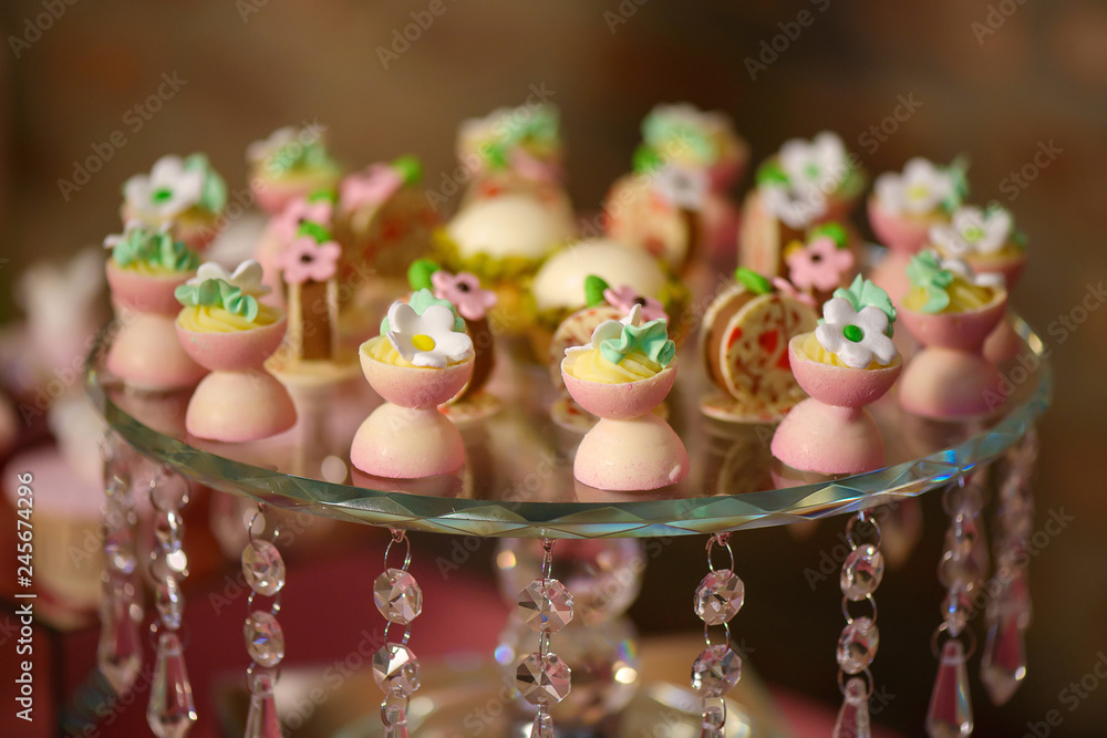 Delicious dessert variety of playful or themed bite-sized cakes shaped as small white and pink chocolate cups with icing sugar flowers, displayed on an embellished crystal cake stand