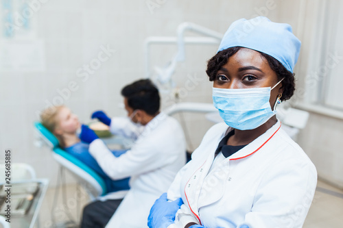 African dentist in mask looks into camera against the background of a doctor treats teeth