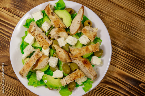 Tasty salad with fried chicken breast, green olives, feta cheese, avocado, lettuce and olive oil on wooden table. Top view