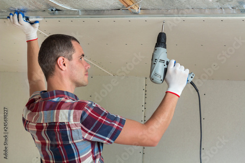 Young man in usual clothing and work gloves fixing drywall suspended ceiling to metal frame using electrical screwdriver on ceiling insulated with shiny aluminum foil. DIY, do it yourself concept. photo