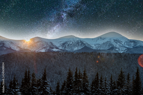 Mountain winter night panorama, Christmas landscape. Steep long ridge mountain peaks covered with snow and dense spruce forest hills, lit by setting sun horizon on dark blue starry sky background.