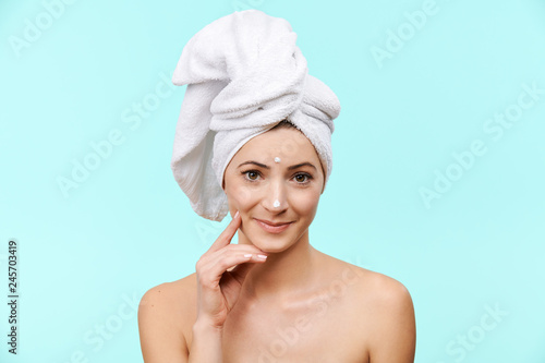 Smiling mid 30s woman with moisturizing cream on her face. Photo of attractive caucasian woman with healthy skin isolated on pastel blue background. Beauty and Skincare concept.
