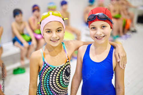 Portrait of two smiling little girls in swimsuit standing and embracing during swimming lesson in pool