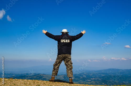 Man with spread arms in a joyful pose, on a mountain top, watching idyllic landscape of the mountain. Translation of the text on the jacket: "Serbia"