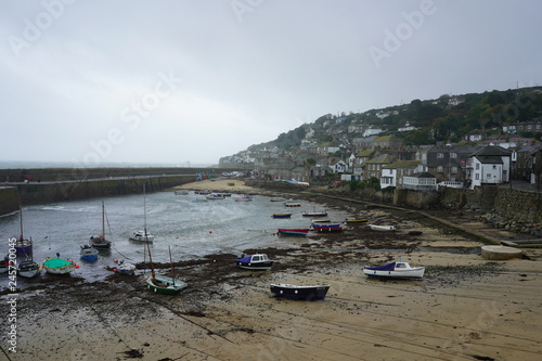 Boats on the beach behind the port walls and the village houses, grey cloudy day. Mousehole, Cornwall, UK