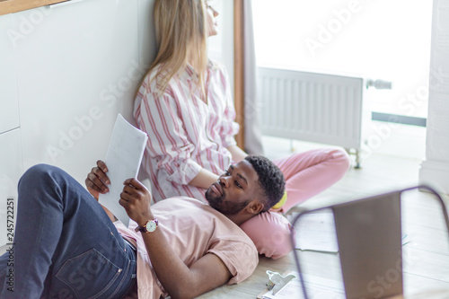 African man reading papers while resting on knees of his blonde long-haired caucasian girlfriend, sitting in sleepwear on the floor at home